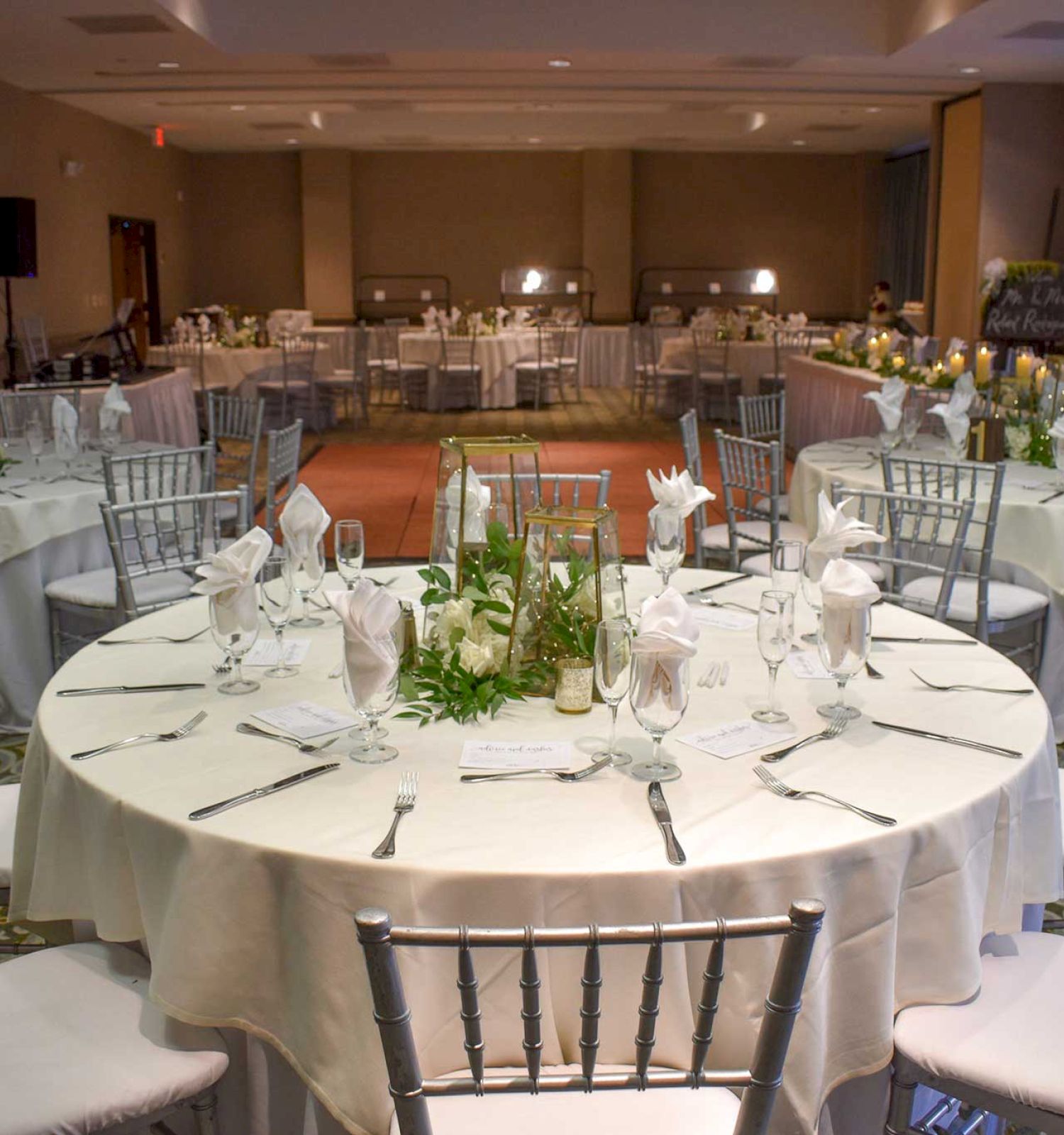 A decorated banquet hall with round tables set with white tablecloths, chairs, glassware, and floral centerpieces, ready for a formal event.