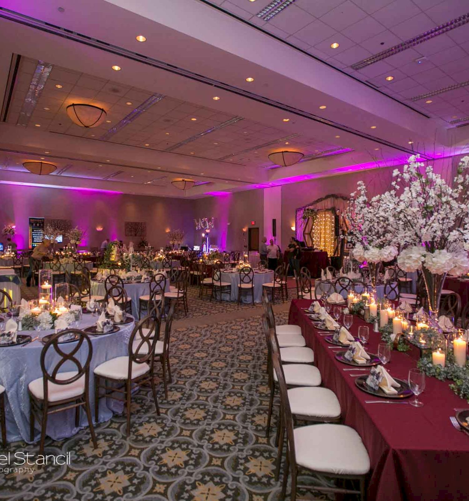 The image shows an elegantly decorated banquet hall with round and long tables adorned with flowers and candles, set up for an event.