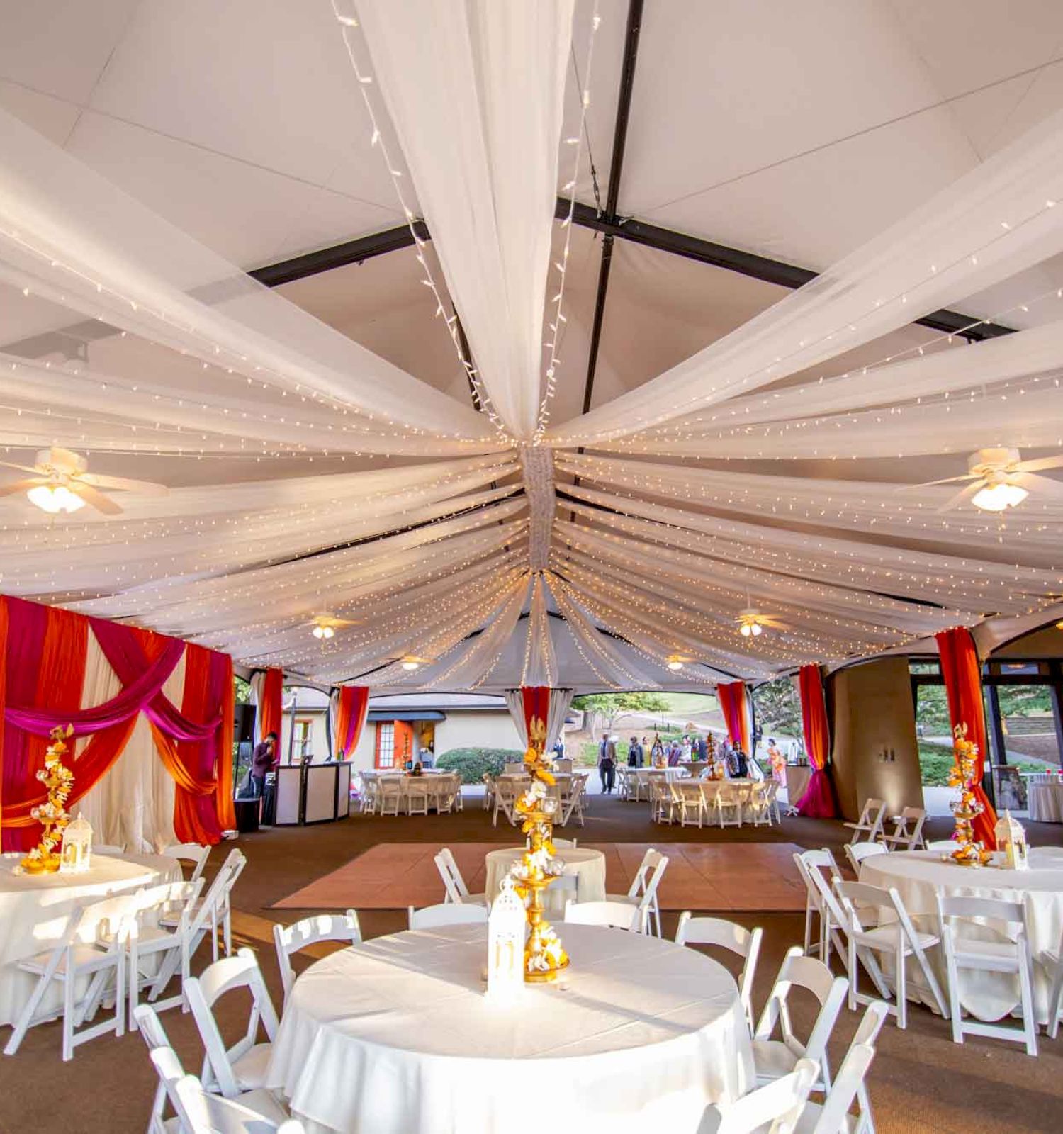 The image shows a decorated venue with white draped ceiling fabric, white tables and chairs, and golden centerpieces, likely set up for a wedding or event.