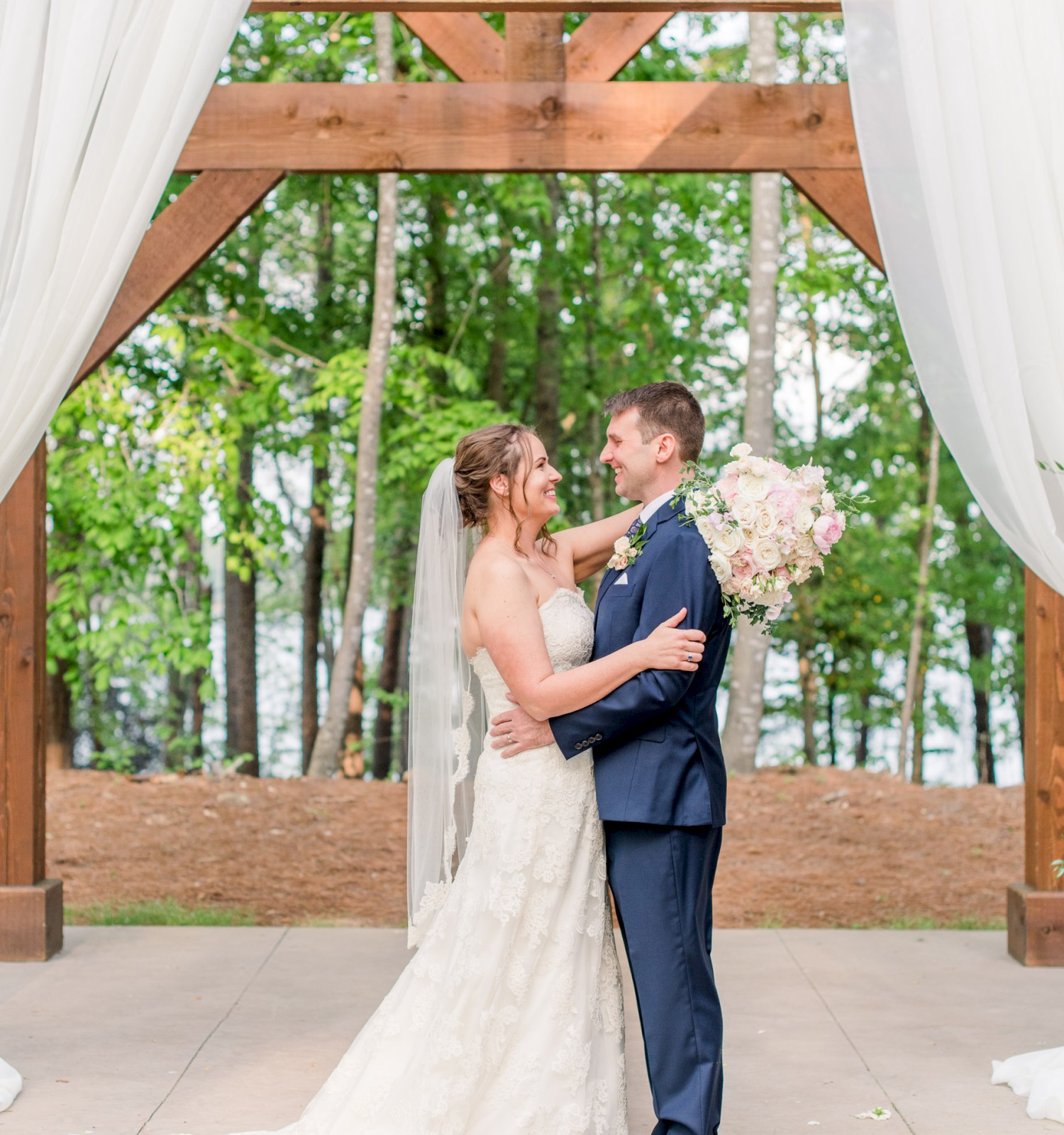 A bride and groom are embracing under a wooden arch with white drapes in an outdoor setting, likely during their wedding ceremony.