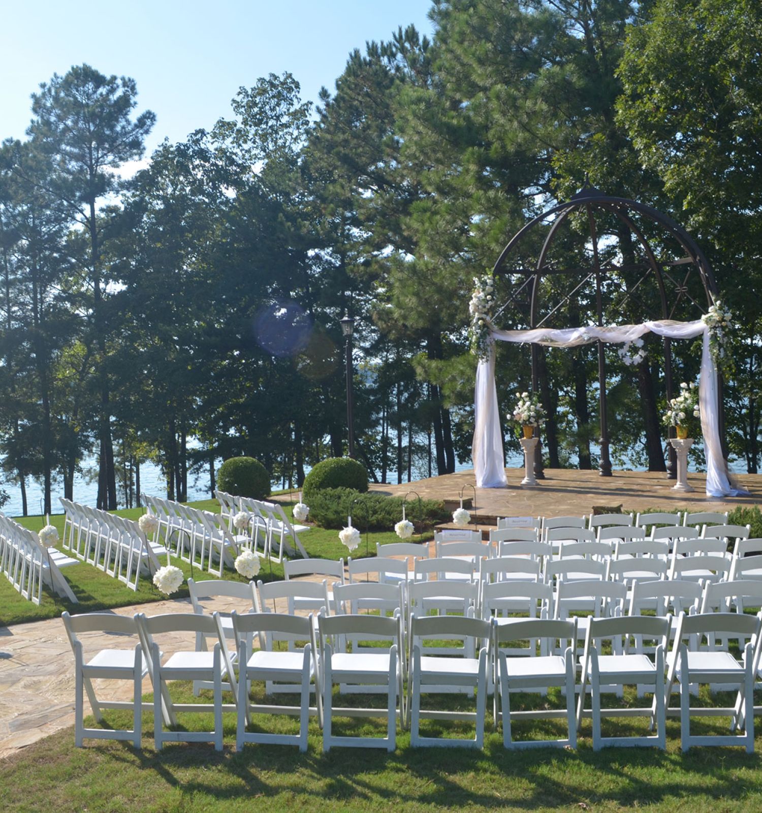 An outdoor wedding setup with white chairs, a decorated arch, and a scenic background of trees and a body of water.
