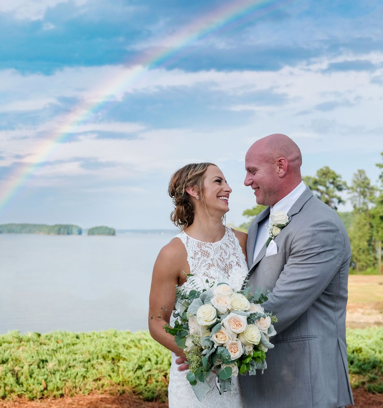 A couple in wedding attire stands by a lake with a rainbow in the background, smiling at each other while holding a bouquet of white roses.