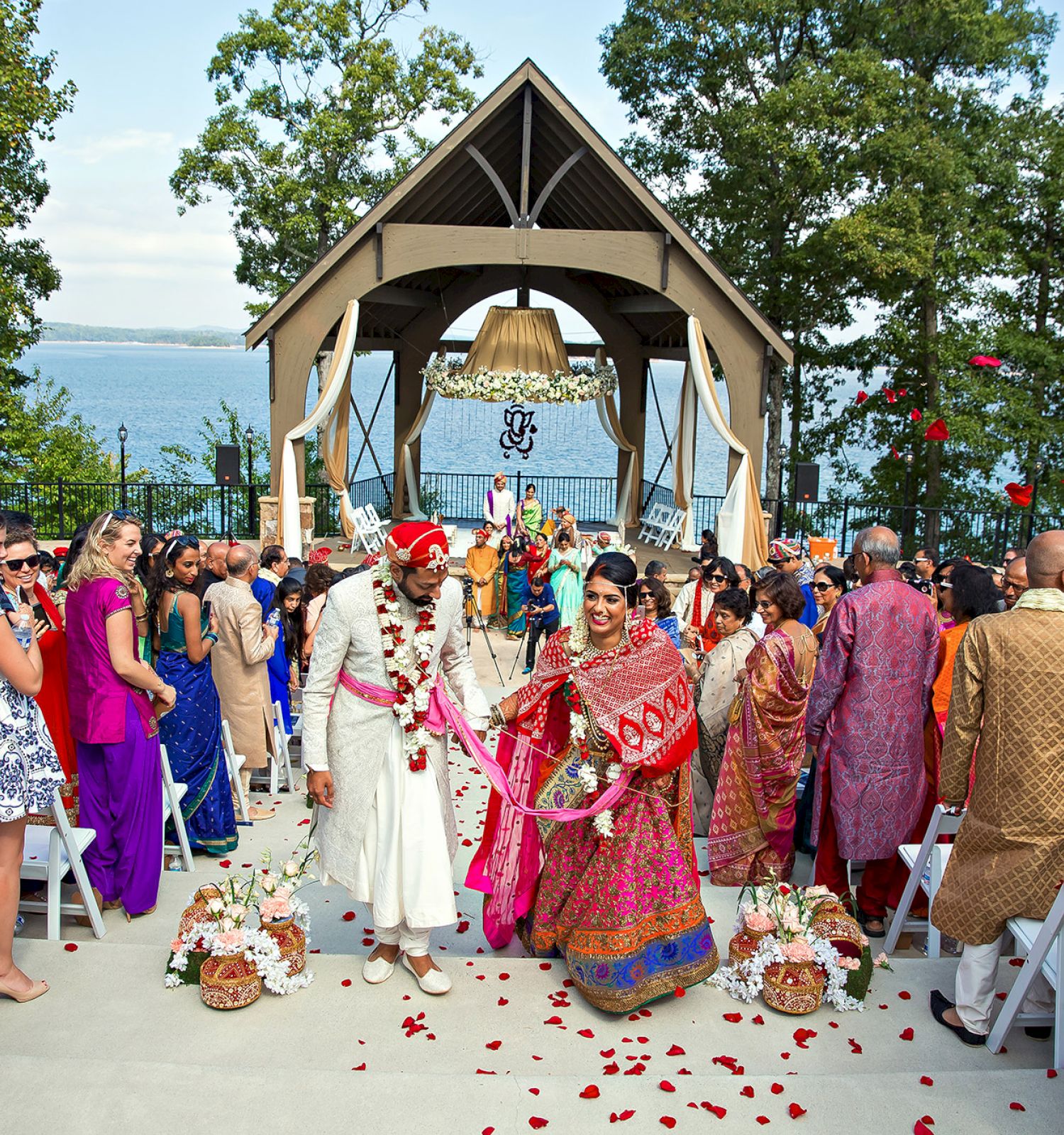 A couple in traditional attire is having an outdoor wedding ceremony by a lake, surrounded by guests and decorated with flowers.