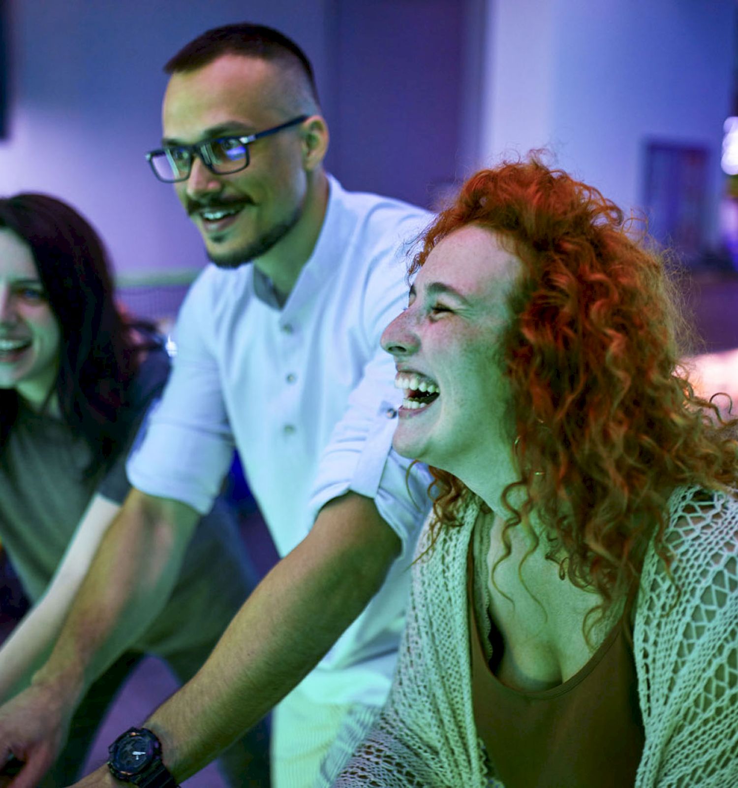 A group of four friends is enjoying playing arcade games, smiling and laughing together in a brightly lit gaming area at night.