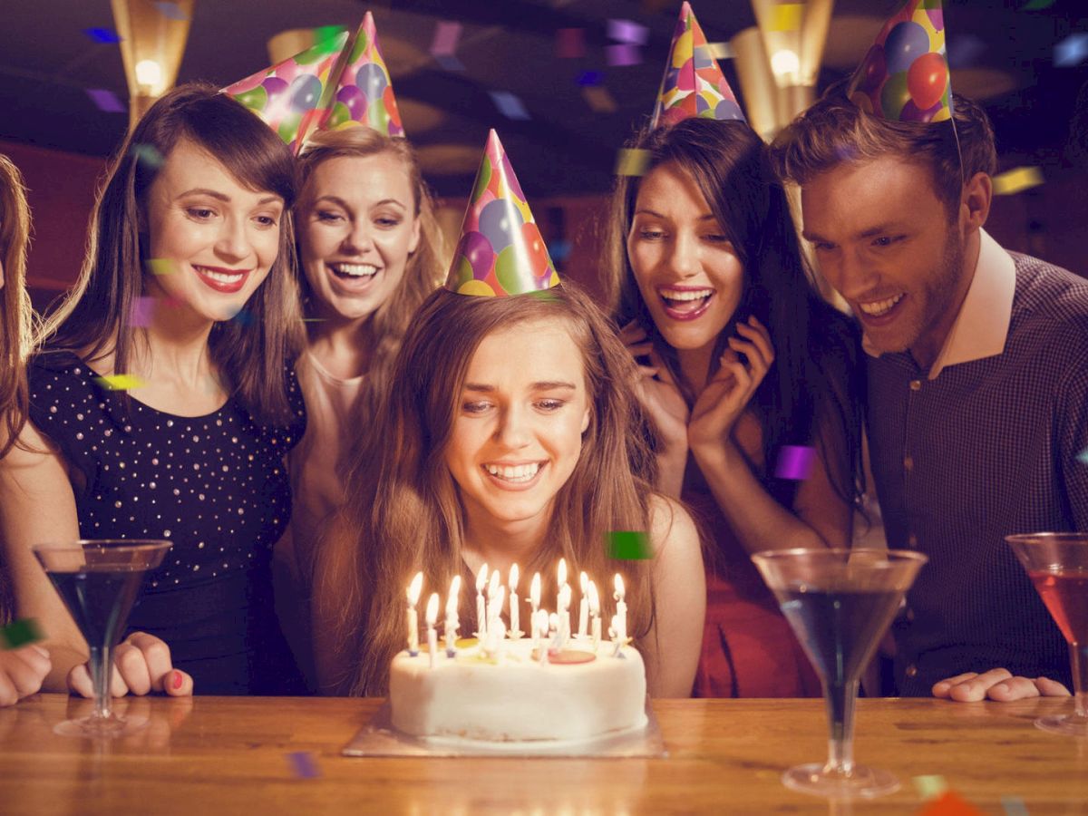 A group of seven people celebrating a birthday with party hats and drinks. A woman in the center is about to blow out candles on a cake.