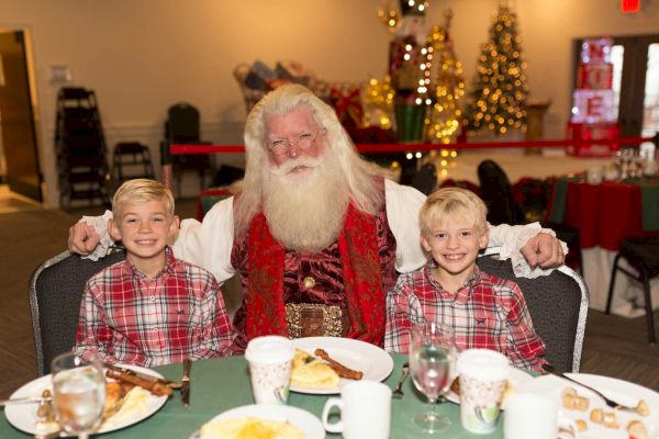 A person dressed as Santa Claus sits with two smiling children at a table, with Christmas decorations in the background.