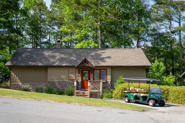A single-story house with a brown roof and beige siding, located in a wooded area. A green golf cart is parked in the driveway at the front.