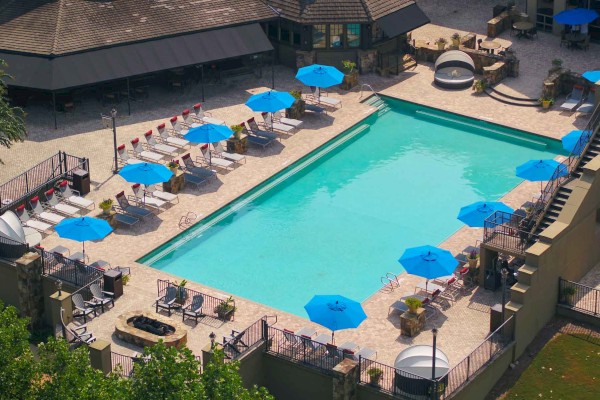 An outdoor pool area with blue umbrellas, lounge chairs, tables, and a surrounding deck, near a building with a roof.