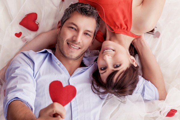 A couple lying down, smiling at the camera, holding a red heart shape. They are surrounded by white fabric and additional red heart shapes.