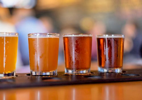 The image shows four glasses of beer on a wooden tray, each containing different types of beer with varying colors and opacity.