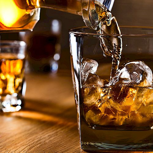 A glass with ice being filled with amber-colored liquor from a decanter, with two more filled glasses in the background on a wooden surface.