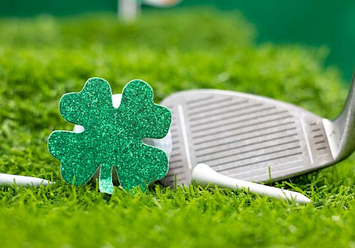 A glittery green shamrock ornament, white golf tees, and a golf club are situated on vibrant green grass.