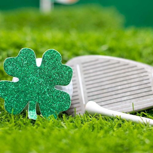 A glittery green shamrock ornament, white golf tees, and a golf club are situated on vibrant green grass.