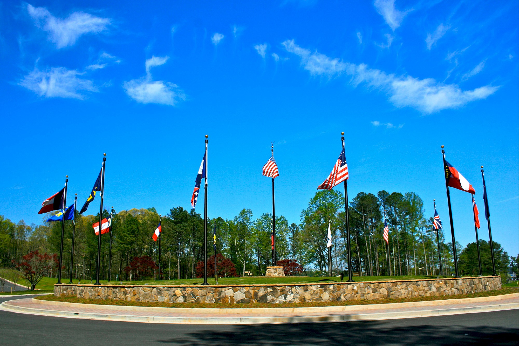 A roundabout with multiple flagpoles displaying various national and state flags set against a bright blue sky with scattered clouds.
