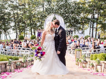 A bride and groom share a kiss at an outdoor wedding ceremony, surrounded by guests seated under a canopy of trees, with petals and flowers adorning the aisle.