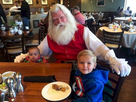 A person dressed as Santa Claus poses with two children at a restaurant, with food and table settings visible around them, creating a festive atmosphere.