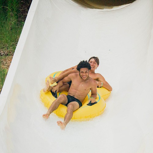 Two people riding down a white water slide on a yellow inflatable raft, smiling and enjoying the experience.