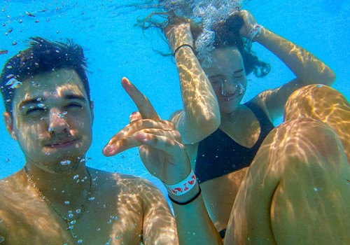 Two people are underwater in a pool. One is making a hand gesture while the other adjusts their hair. Both appear to be enjoying their time.