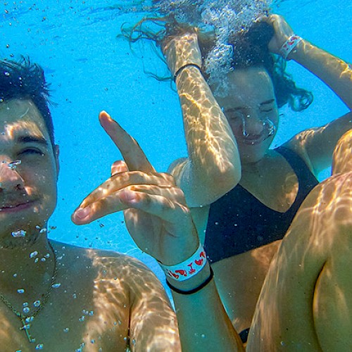 Two people are underwater in a pool. One is making a hand gesture while the other adjusts their hair. Both appear to be enjoying their time.