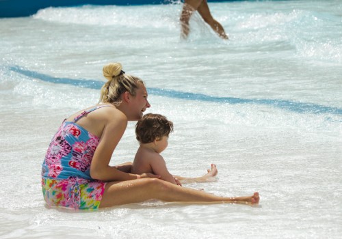 A woman and a child are sitting in shallow water at a beach or water park, enjoying the waves and sunshine. Another person walks by in the background.