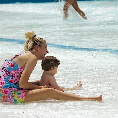 A woman and a child are sitting in shallow water at a beach or water park, enjoying the waves and sunshine. Another person walks by in the background.