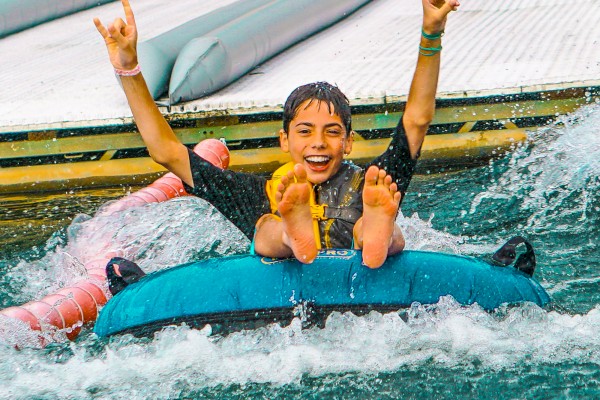 A person joyfully rides an inner tube in water, holding up hands in excitement, while wearing a life jacket.