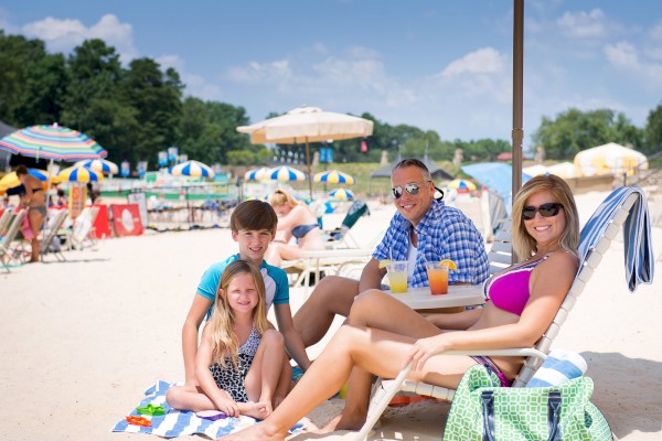 A family of four enjoys a sunny day at the beach, lounging on chairs with drinks, and kids playing on a towel in the sand.