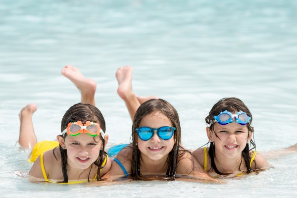 Three kids in swimsuits lying in shallow water, wearing colorful goggles.