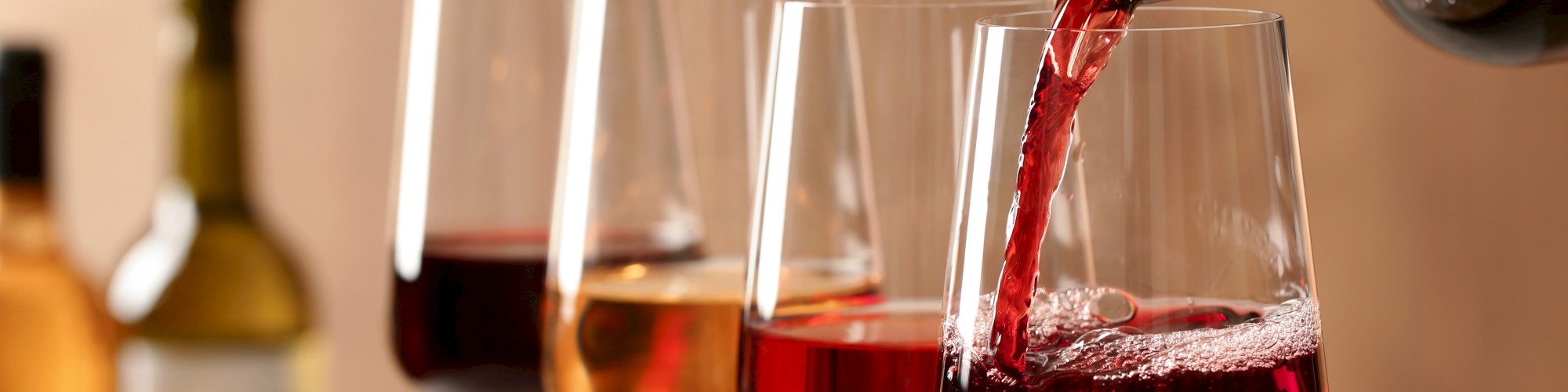A bottle pouring red wine into one of four glasses, with more wine bottles in the background.