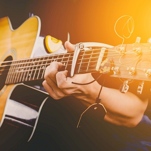 A person is playing an acoustic guitar, with fingers positioned on the strings and frets. The photo has a warm, sunlit ambiance, capturing the moment.