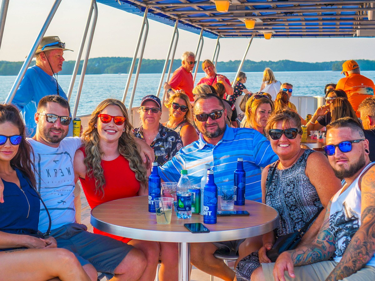 A group of people enjoying a boat trip, seated together with drinks on a sunny day. They are all smiling and wearing sunglasses.