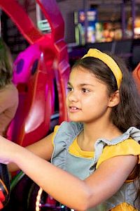 A young girl wearing a headband and dress is focused while playing an arcade game, sitting in front of a pink machine.
