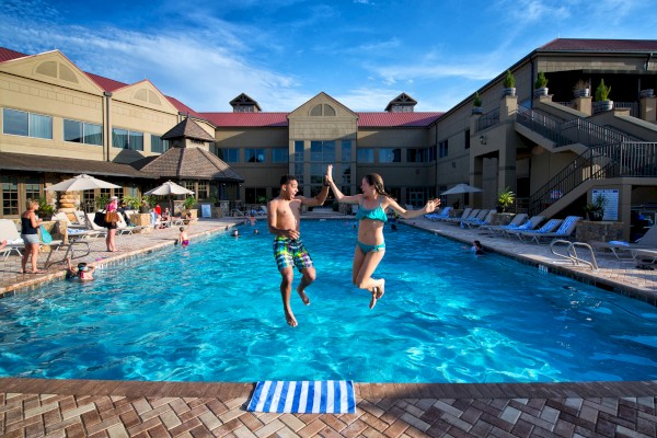 Two people are jumping into a large outdoor pool at a resort. Other guests are relaxing and swimming. The sky is clear, and the buildings are visible.