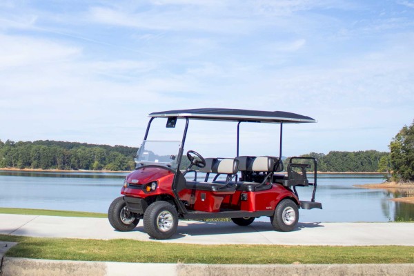 A red, six-seater golf cart is parked on a path near a scenic lake, surrounded by greenery and a partly cloudy sky.