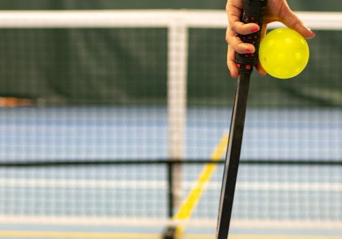 A hand holding a paddle and pickleball in front of a net on a court.