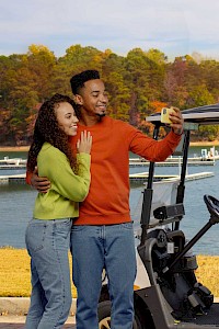 A couple takes a selfie beside a golf cart near a lake with autumn trees in the background.