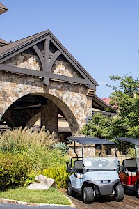 Two golf carts, one blue and one red, are parked in front of a rustic stone building with greenery and trees around it on a clear, sunny day.