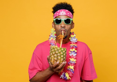 A person wearing a pink shirt, green sunglasses, and a flower lei sips from a pineapple with a straw against a yellow background.