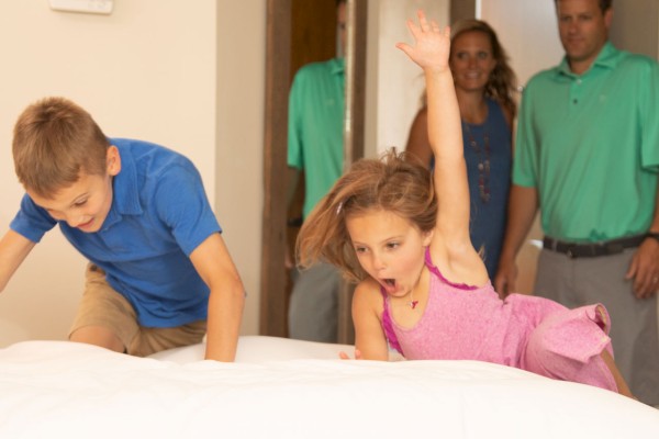 Two children are jumping onto a bed while two adults watch them from the doorway, smiling in the background.
