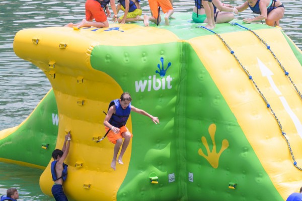 People in life vests are playing on a large inflatable water obstacle with the name 