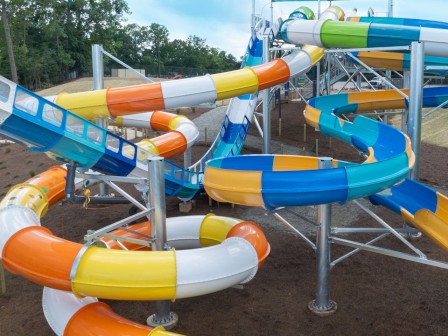 The image features a colorful water slide at an amusement park, with intertwined tubes in orange, yellow, blue, and white, set against a green background.