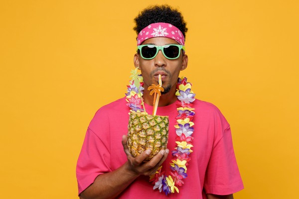 A person in a pink shirt, with sunglasses and a flower lei, sips a drink from a pineapple against a bright yellow background.