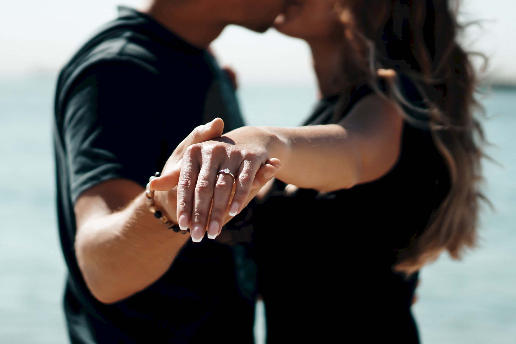 A couple is kissing and holding hands, with a focus on the woman's ring, set against a blurred waterfront background.