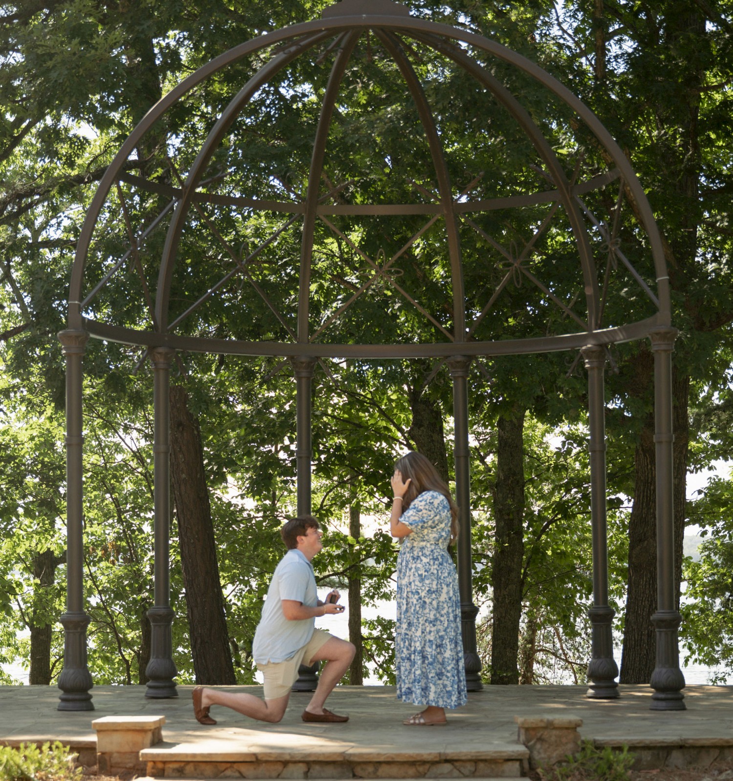 A person is kneeling and proposing to another under a decorative metal gazebo in a lush, forested setting, with a body of water in the background.