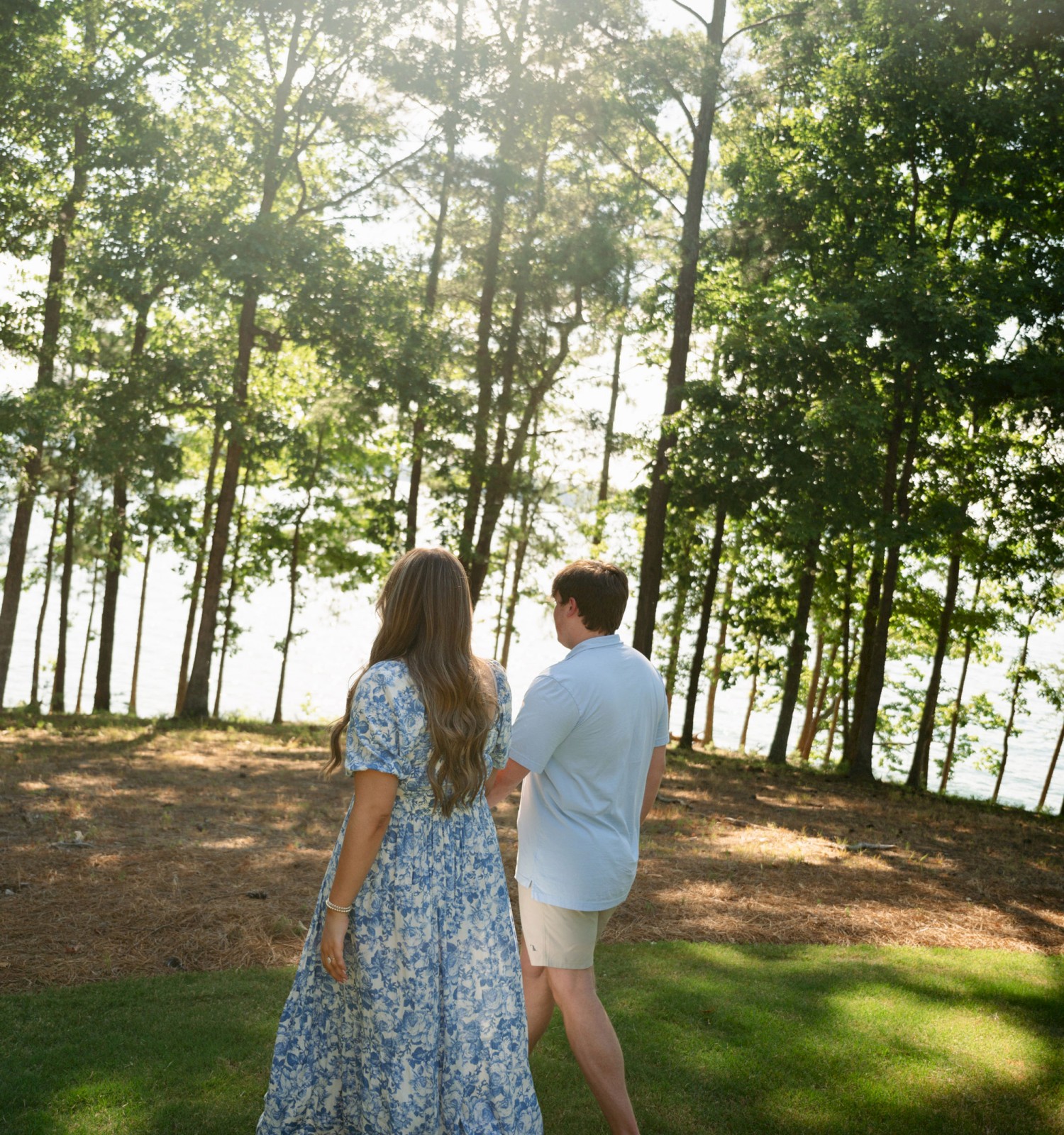 A couple is seen from the back standing on a grassy area surrounded by tall trees with sunlight filtering through the leaves, near a body of water.