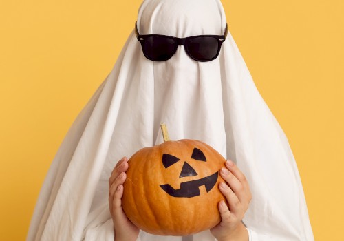 A person dressed as a ghost with sunglasses holds a carved pumpkin against a yellow background.