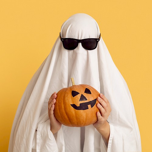 A person dressed as a ghost with sunglasses holds a carved pumpkin against a yellow background.