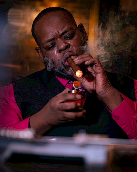 A person in a red shirt lights a cigar in a dimly lit room, with smoke and a relaxed ambiance.