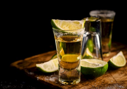 The image shows glasses of golden liquid, probably tequila, garnished with lime wedges and surrounded by additional lime slices, on a wooden board.