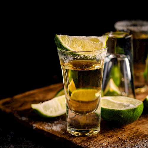 The image shows glasses of golden liquid, probably tequila, garnished with lime wedges and surrounded by additional lime slices, on a wooden board.